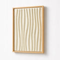 A framed abstract wall art piece featuring vertical wavy ridges in a warm cream or off-white tone, enclosed in a light natural wood frame and mounted on a plain white wall.