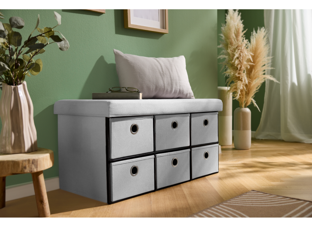 A light gray upholstered bench with six fabric storage drawers underneath, styled in a living room setting with a green wall, wooden floor, and decorative plants.