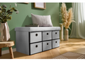 A light gray upholstered bench with six fabric storage drawers underneath, styled in a living room setting with a green wall, wooden floor, and decorative plants.